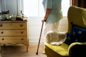 A picture of an elderly woman standing, holding a cane in an adult foster care room. A green chair and a wood dresser.
